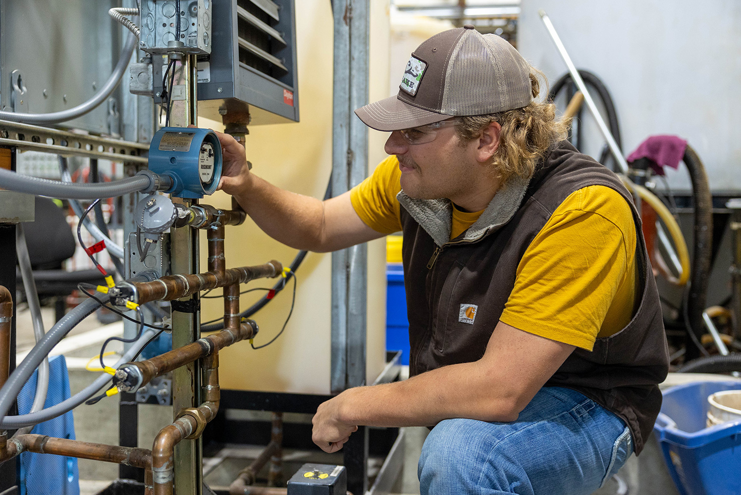 man with brown snapback hat and yellow t-shirt inspecting pipes and a gauge