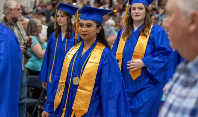 Graduates wearing blue caps and gowns with gold stoles walk in a commencement ceremony. One graduate in the foreground wears honor cords and a medallion, holding a program. Family and friends are seated in the background, watching and taking photos.