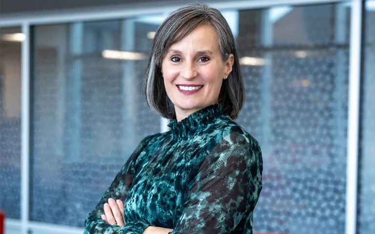 woman with short dark gray and brown hair with a teal and black turtleneck shirt, crossing her arms in front of windows