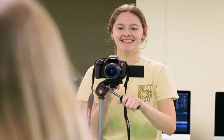 girl with dark hair behind a camera smiling