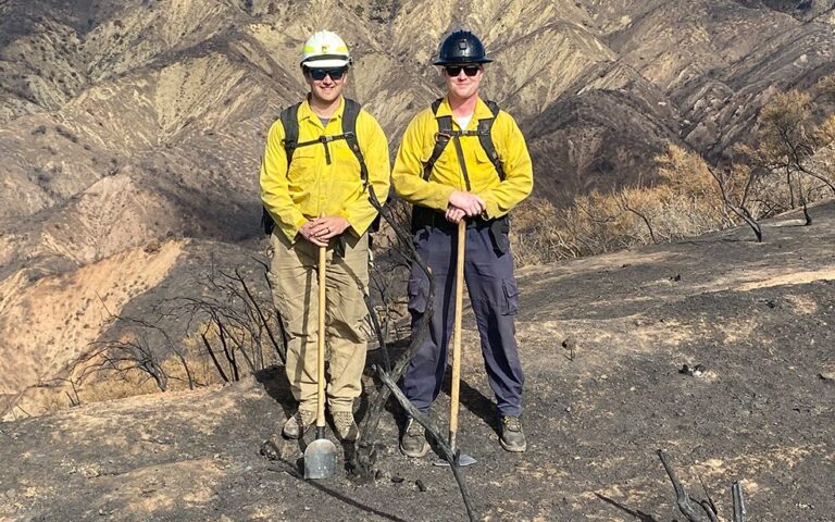 Two firefighters in yellow protective gear and helmets stand side by side on a scorched hillside with charred grass around them. They each hold tools and wear backpacks, with barren, burnt mountains visible in the background.