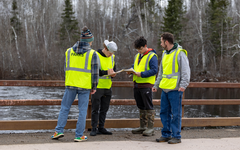 four male students outside on a bridge with clipboards and yellow safety vests on calculating water quality