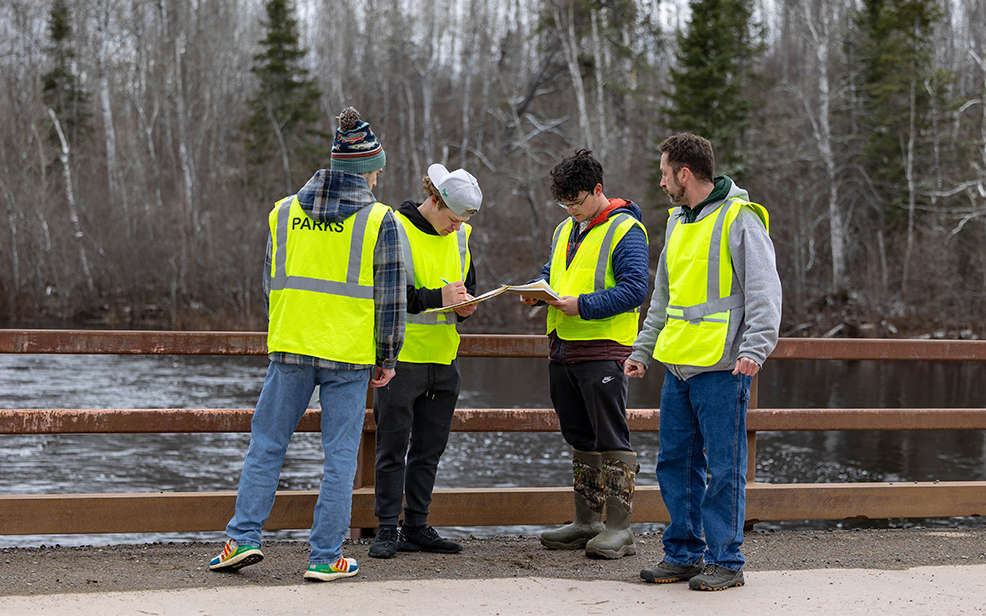 four male students outside on a bridge with clipboards and yellow safety vests on calculating water quality