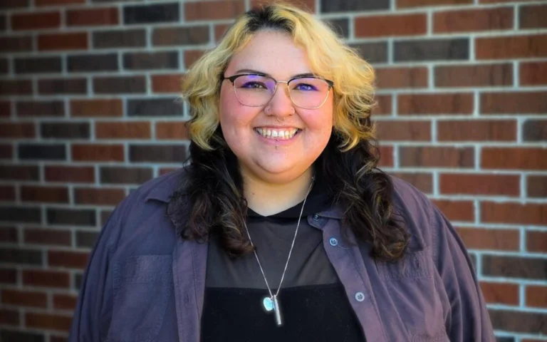 woman with blonde hair on the top, and dark brown underneath, smiling with a purple flannel shirt and black t shirt underneath.