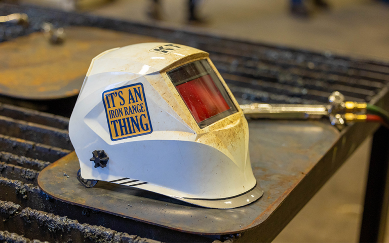 white welding helmet with a yellow sticker with blue writing reading "it's an iron range thing". the welding helmet is on top of a piece of sheet metal