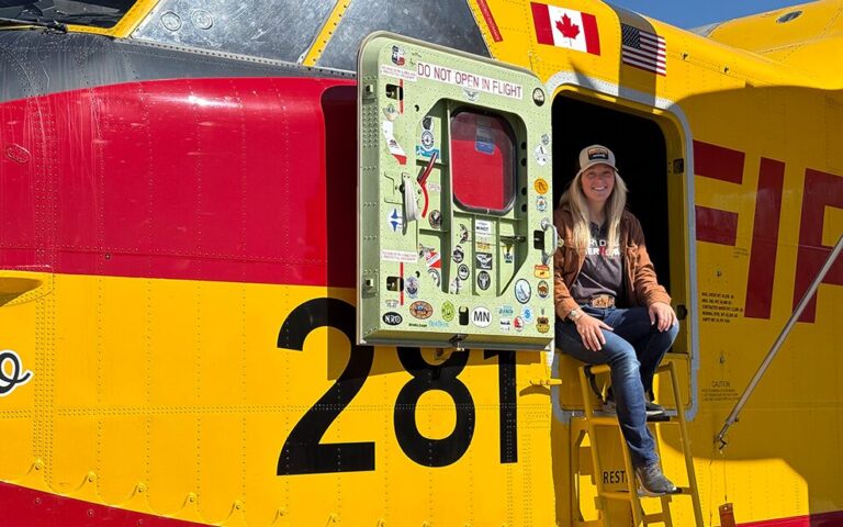 caucasian woman with blonde hair in a yellow fire fighting plane smiling with blue jeans and a gray shirt and a trucker hat