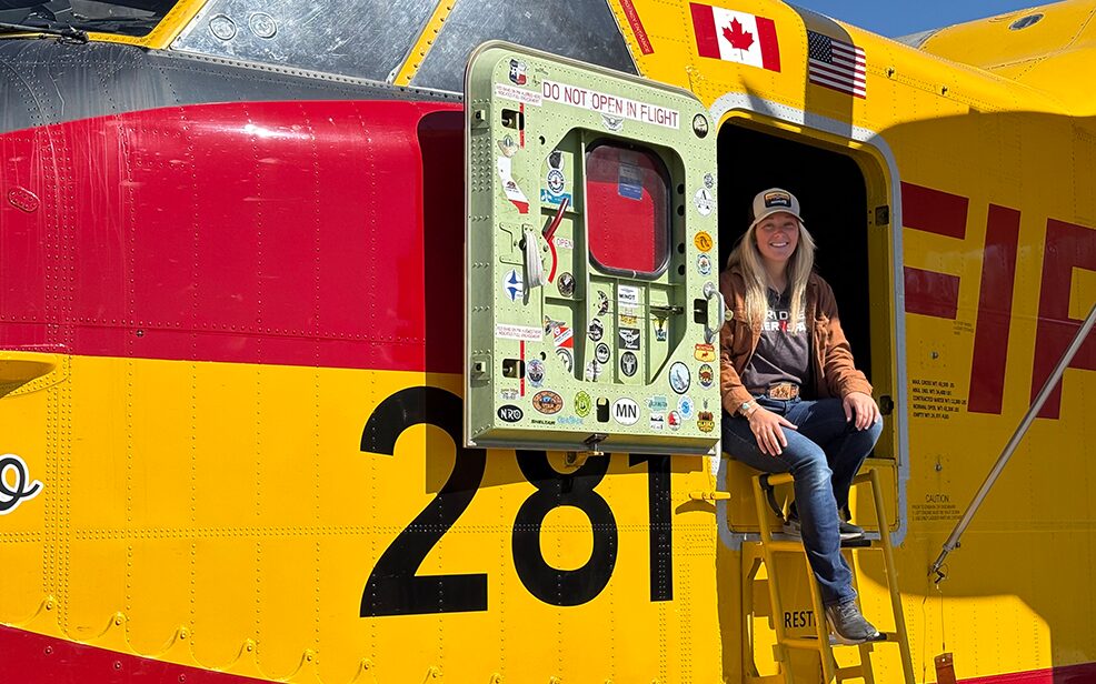 caucasian woman with blonde hair in a yellow fire fighting plane smiling with blue jeans and a gray shirt and a trucker hat