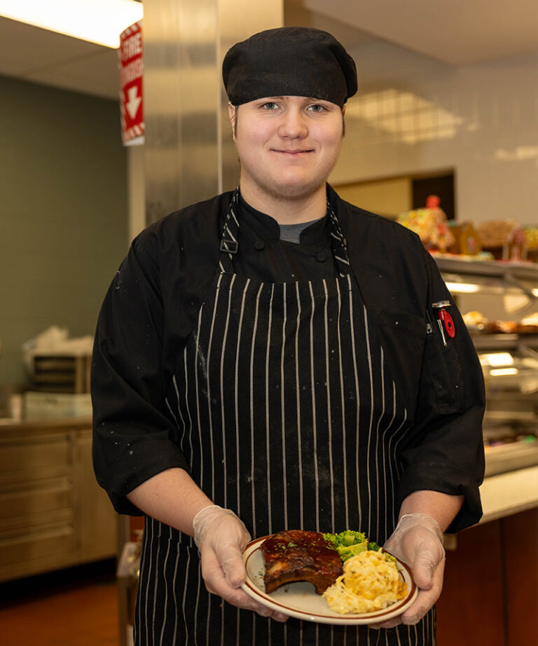 A student chef in a black uniform and striped apron holding a plate with ribs, mashed potatoes, and green vegetables in a commercial kitchen setting