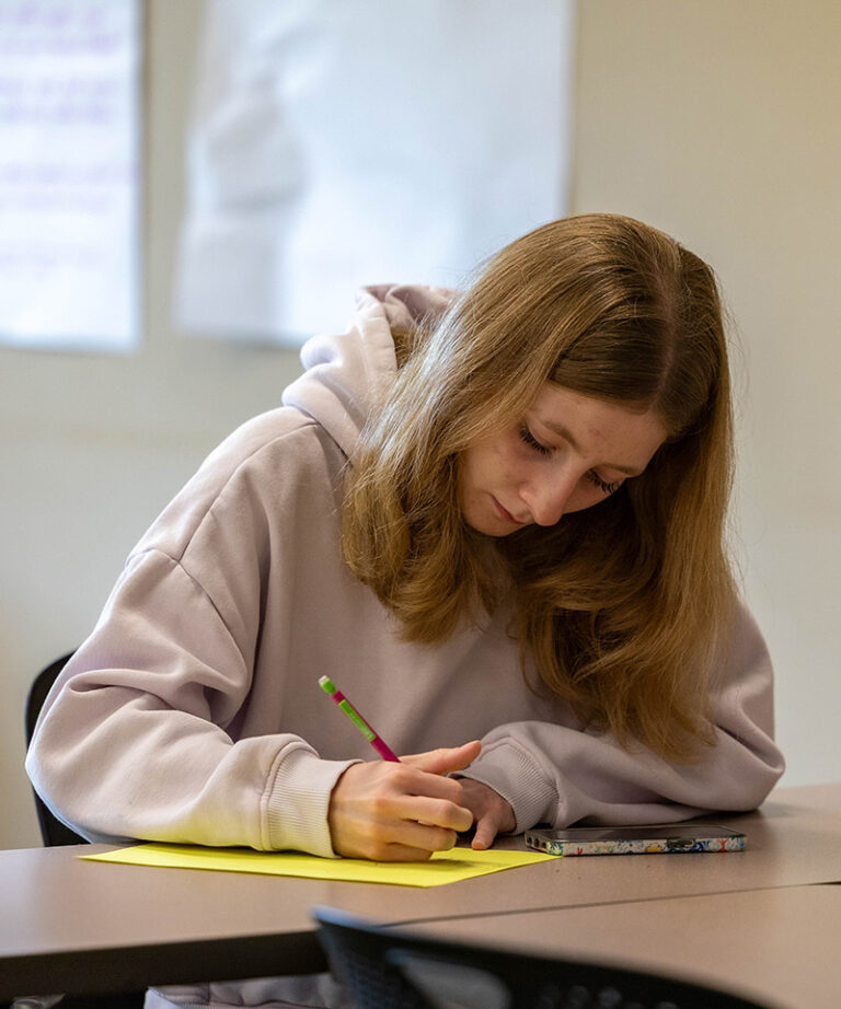 female student with light blonde hair wearing a tan sweatshirt writing on a yellow piece of paper while sitting at a table