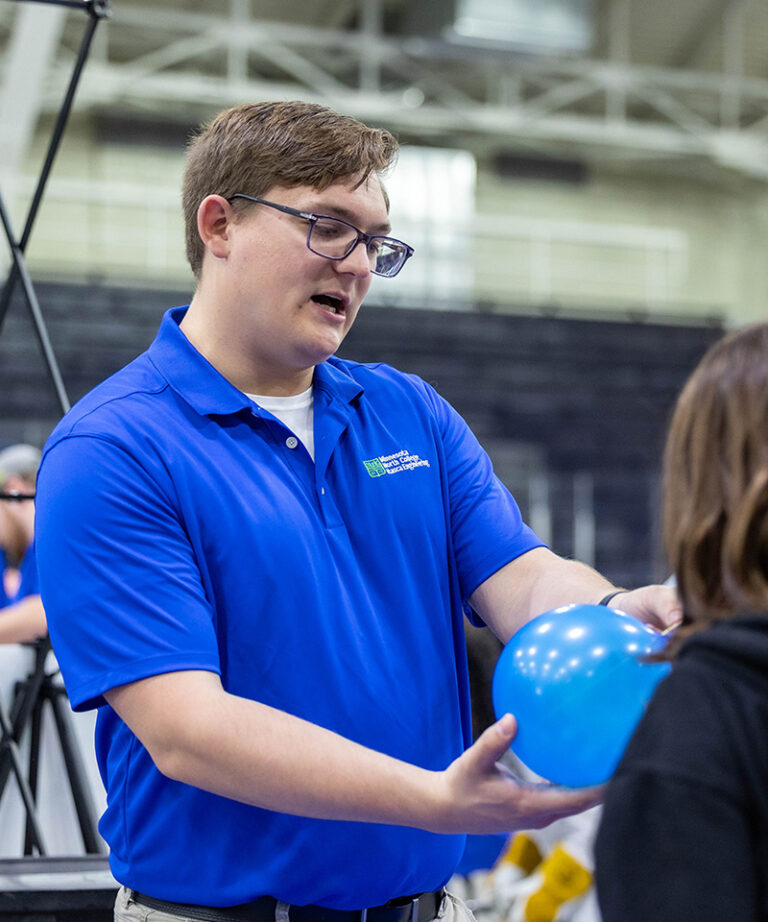male student with glasses wearing a blue polo holding a ballon showing another student