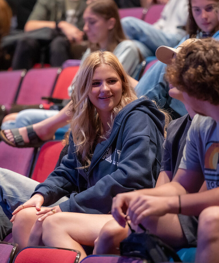 blonde female student sitting in a theatre smiling while talking to another person