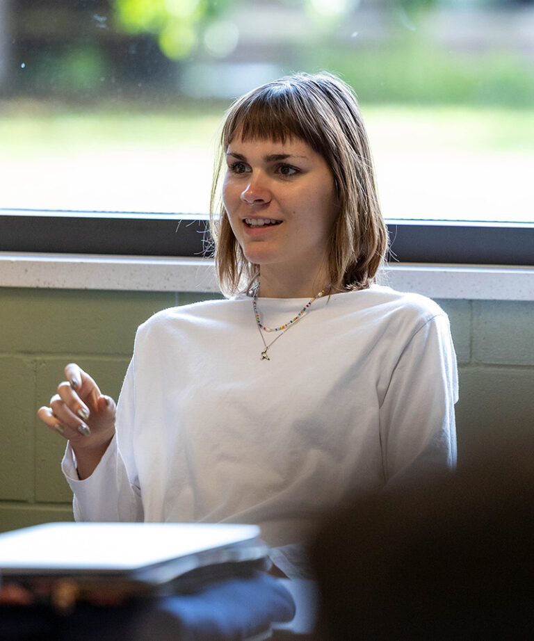 caucasian white female student with shoulder length hair and bangs wearing a white shirt sitting in a classroom talking