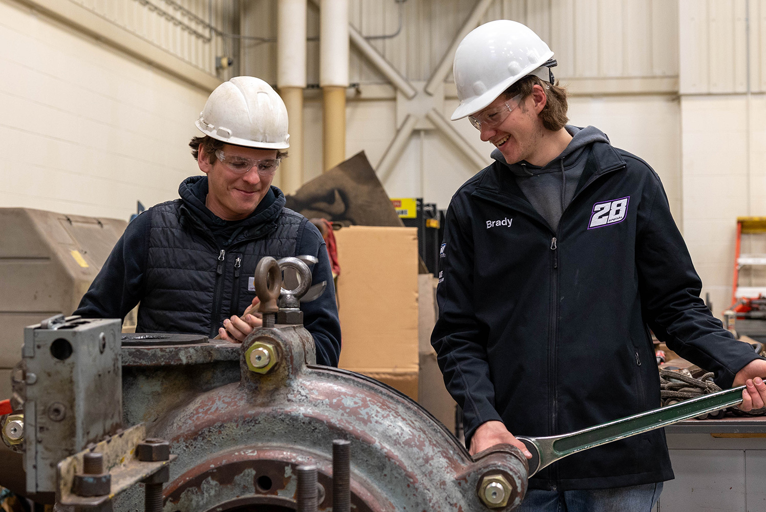 two male students with hard hats standing behind a metal wheel working on it