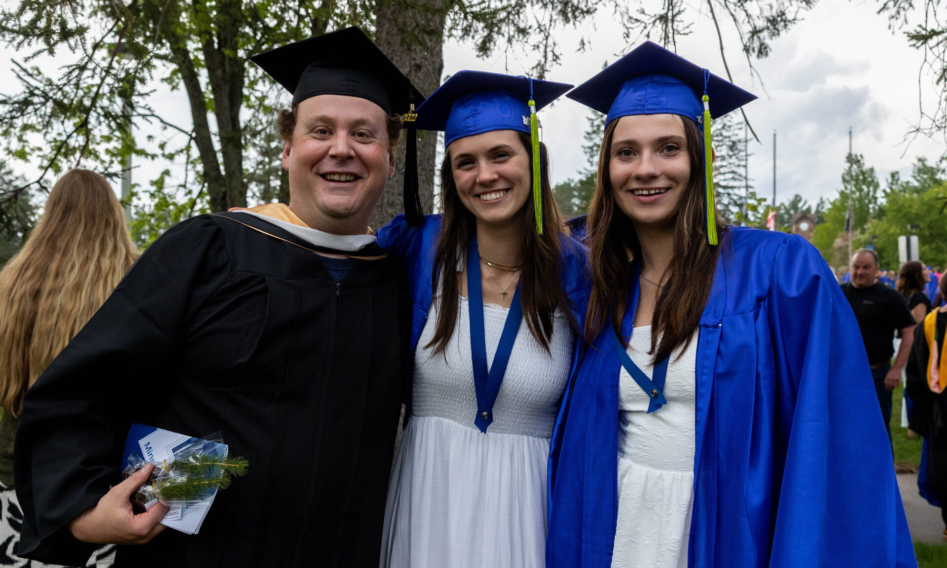 two students and one faculty dressed in caps and gowns for graduation. the male teacher on the left is wearing a black cap and gown, the two female students are wearing white dresses, blue cap and gowns, and are smiling at the camera.