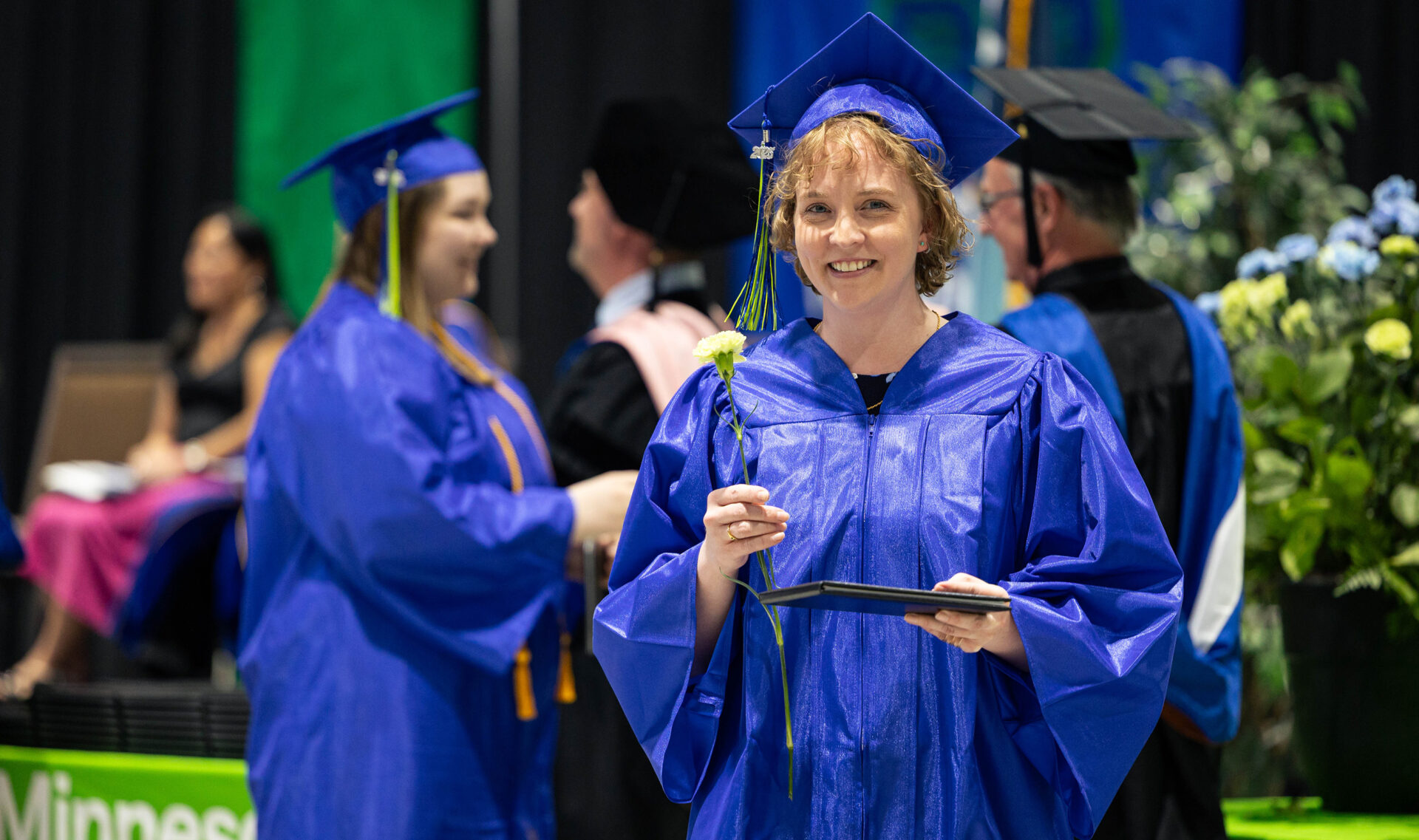 female student at graduation smiling holding diploma and flower up