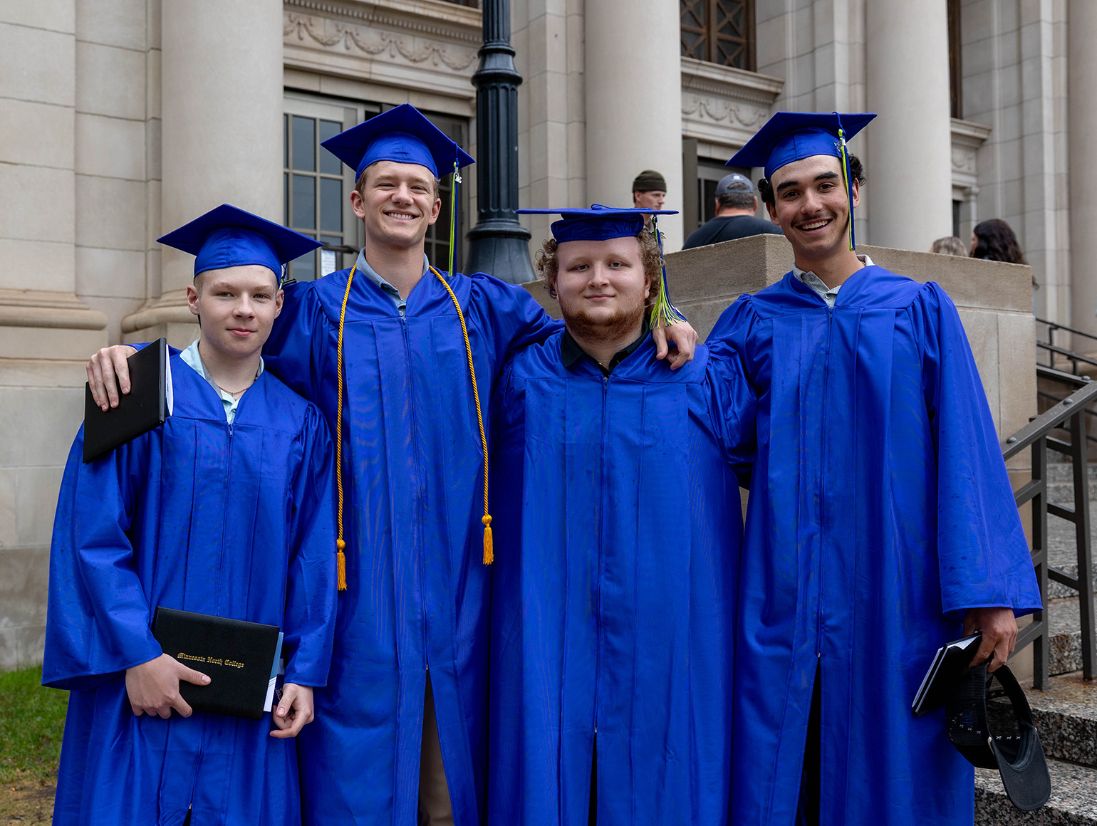 four male students, varying in height, smiling at the camera in blue graduation regalia