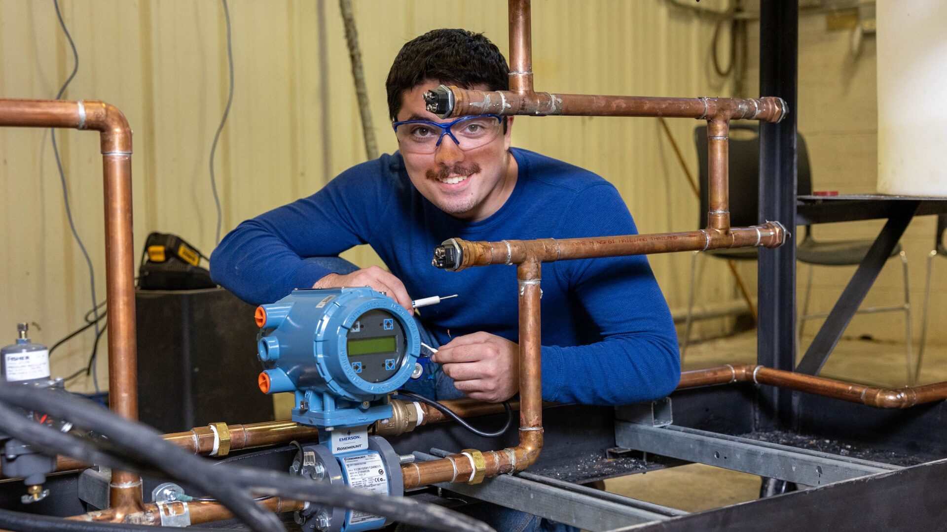 male student squatting behind some pipes and a meter, smiling at the camera